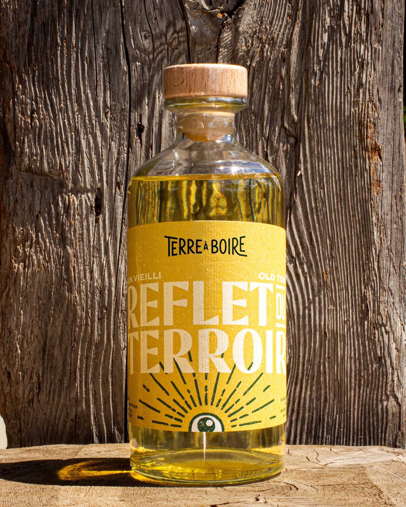 Bouteille de Gin Vieilli Old Tom de Terre à Boire, ornée de l'étiquette jaune « Reflet du Terroir », posée au soleil sur un fond de bois rustique.