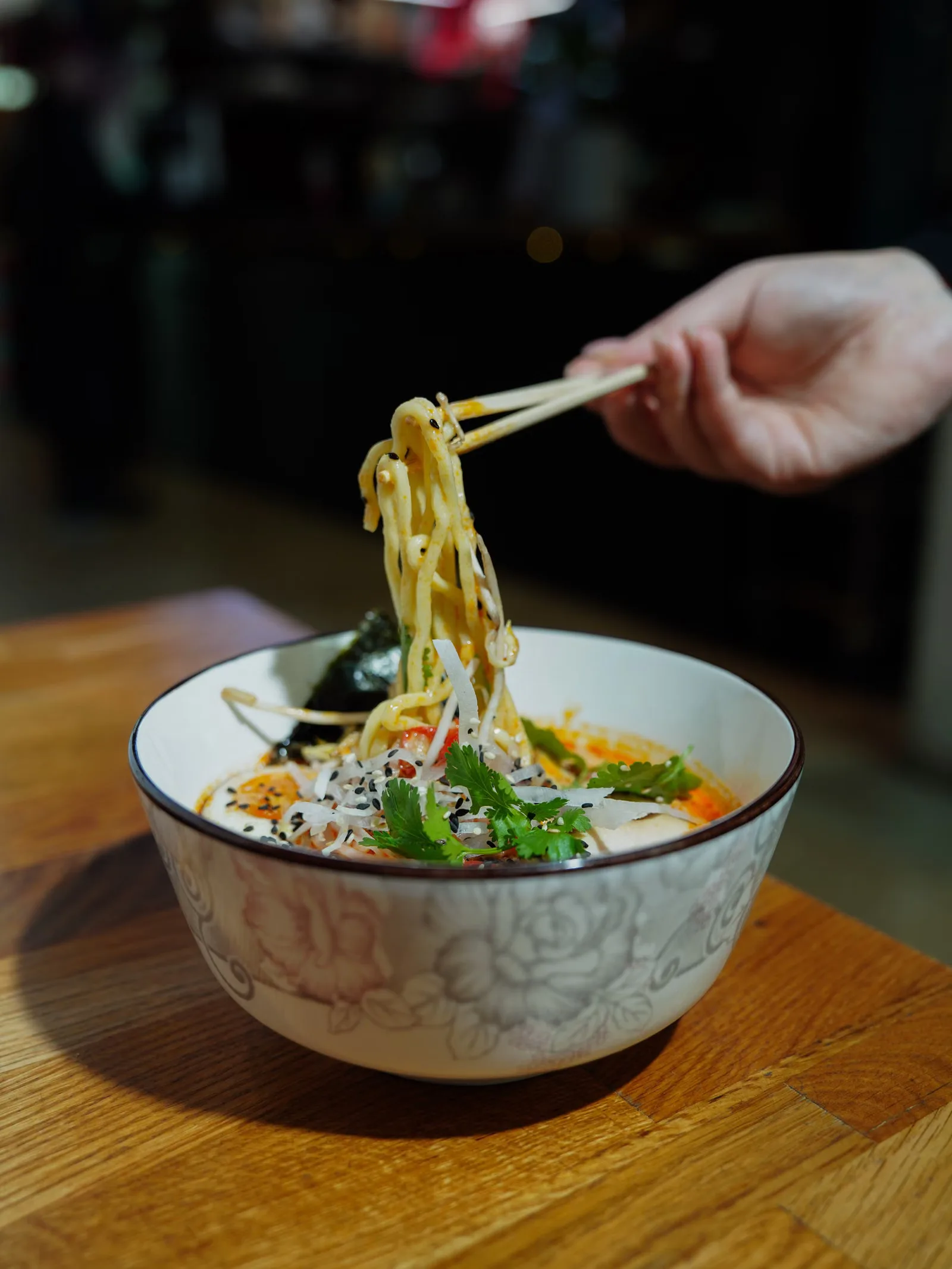 Une main utilisant des baguettes pour soulever des nouilles ramen d'un bol dans un cadre de restaurant.