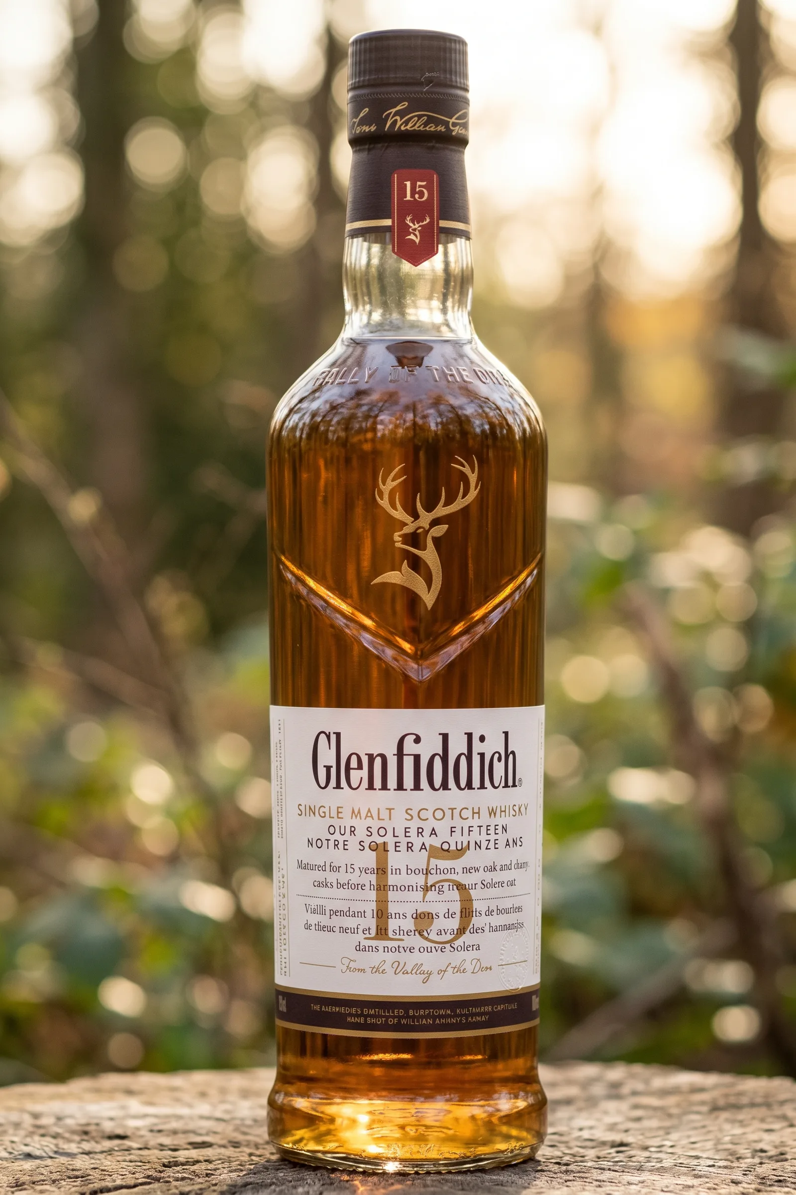 Bouteille de scotch whisky single malt Glenfiddich 15 ans Solera posée sur une surface en bois texturée, photographiée en plein air. La lumière chaude du soleil traverse la bouteille, révélant la riche couleur ambrée du liquide. L'arrière-plan est un sous-bois naturel avec un effet de flou artistiqu